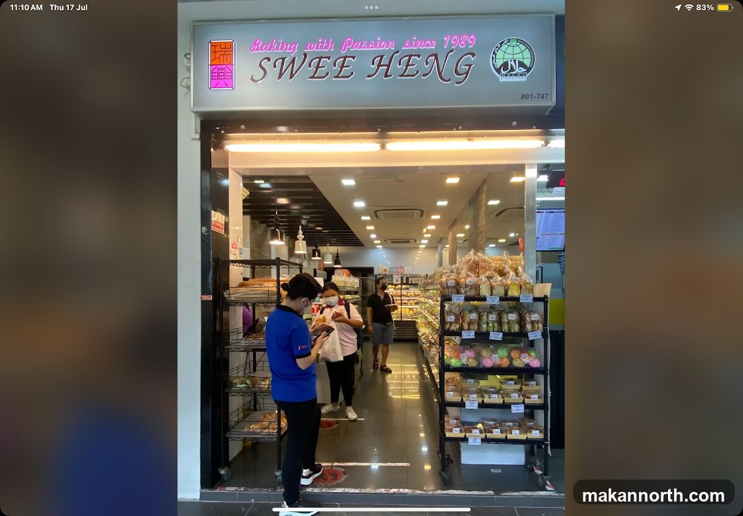 The exterior of a bakery with a prominent sign that reads "Baking with Passion since 1989 SWEE HENG". The entrance is open, revealing shelves stocked with various baked goods. Several people are visible inside and near the entrance.