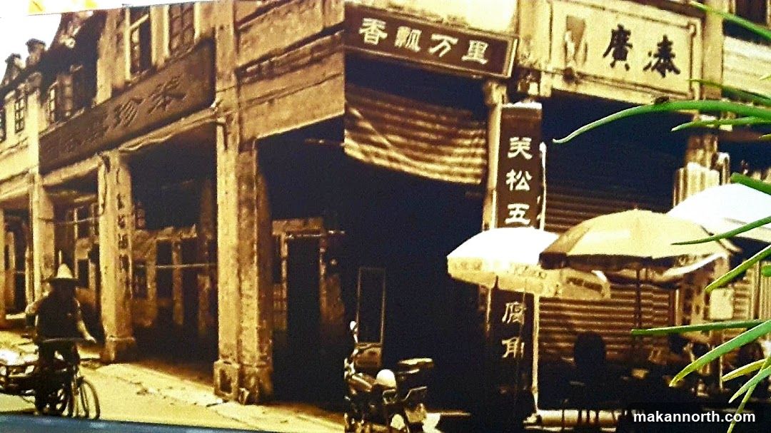 A sepia-toned photograph depicts a street scene with traditional architecture. Several storefronts with ornate facades line the street. One storefront has a sign with Chinese characters above its entrance. Another sign with Chinese characters hangs vertically. A person on a bicycle with a cart is visible on the left side of the image. In the foreground, there are umbrellas and a motorcycle, suggesting an outdoor market or seating area. Green foliage is visible in the upper right corner.