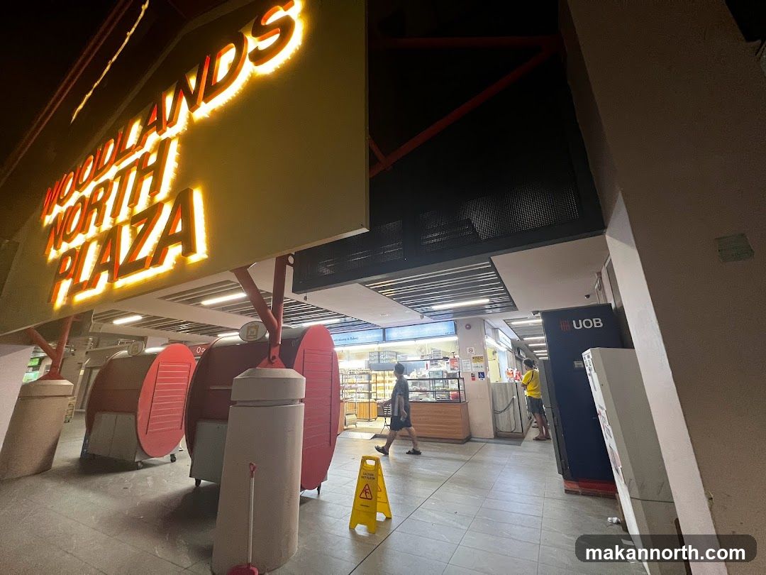 A brightly lit sign for Woodlands North Plaza is visible above an outdoor walkway. In the background, there are several small kiosks and a UOB ATM. People are walking through the area.