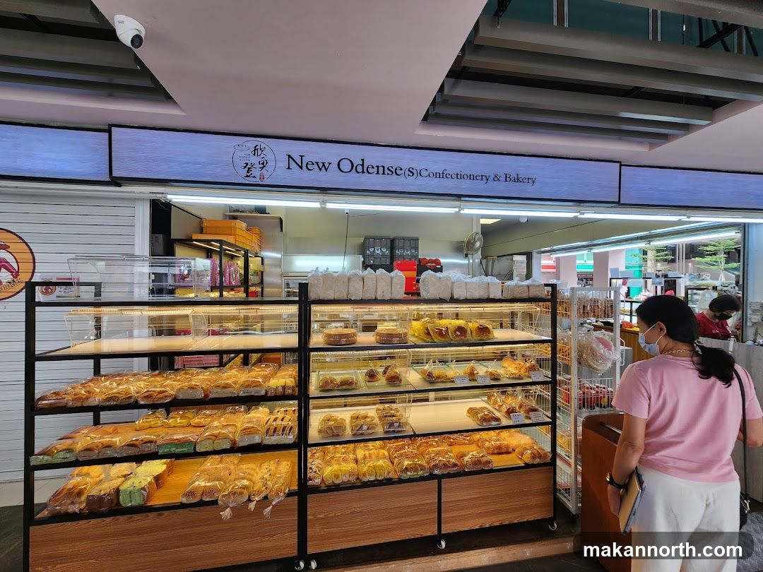 A bakery storefront with a sign that reads "New Odense (S) Confectionery & Bakery". Inside, shelves are filled with various baked goods, including bread, pastries, and cakes, all individually packaged. Two women are visible inside the shop, one browsing the selection.