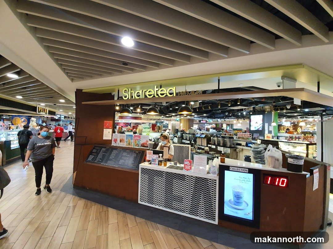 A Sharetea shop front with a menu board and a digital display showing a drink. The shop is located in a mall with other businesses visible in the background.