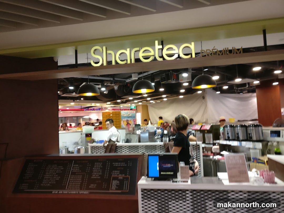 The exterior of a Sharetea shop with the brand name visible above the entrance. Inside, staff are visible behind a counter, and there are various beverage dispensers and equipment. A menu board is displayed prominently in the foreground.