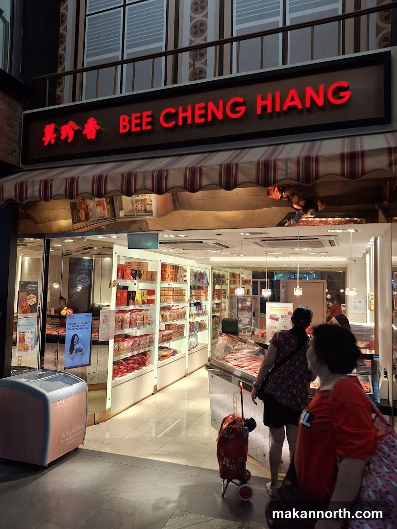 The exterior of a Bee Cheng Hiang shop. The shop has a large red neon sign with the name "BEE CHENG HIANG" in English and Chinese characters. Below the sign is a striped awning. The shop interior is visible, displaying shelves stocked with packaged food items and refrigerated display cases with meat products. Several customers are inside and near the entrance.