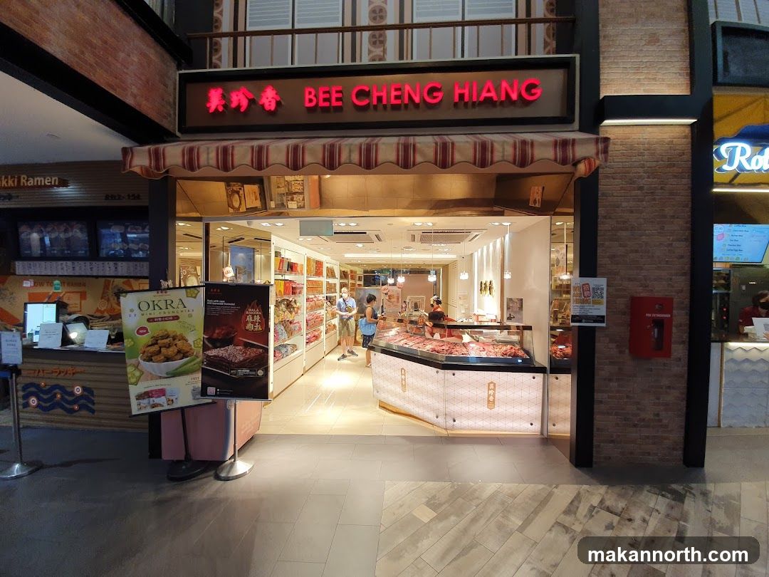 The exterior of a Bee Cheng Hiang shop with its name displayed in red neon letters above the entrance. Inside, shelves are stocked with products, and a display counter showcases meat products. Two people are visible inside the shop. In front of the shop, there are two promotional posters, one for "OKRA EZ Mini Munchies" and another for "WALA WALA".