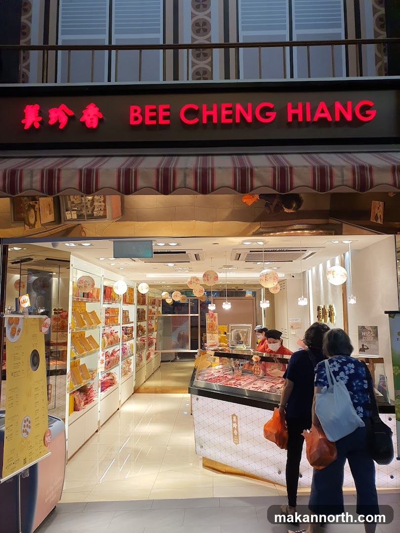 The exterior of a Bee Cheng Hiang shop. The shop has a bright red neon sign with the name in English and Chinese characters. Below the sign is a striped awning. The storefront is open, revealing shelves stocked with packaged goods and a display case with meat. Two customers are standing at the counter, and a staff member is behind it.
