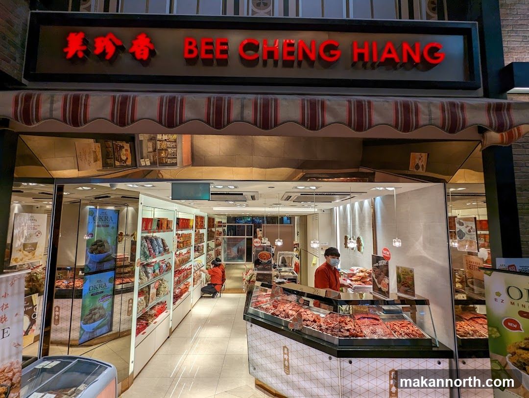 The exterior of a Bee Cheng Hiang shop. The shop has a large red neon sign with the name "BEE CHENG HIANG" in English and Chinese characters. Below the sign is a striped awning. The storefront is open, revealing a well-lit interior with shelves stocked with various food products. A display case in the foreground showcases rows of dried meat. Two employees are visible inside the shop, one attending to the display case and another stocking shelves.