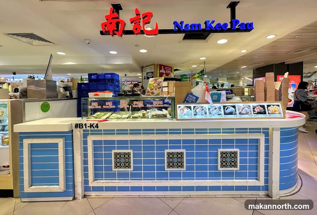 A food stall with a blue tiled counter and a sign that says "Nam Kee Pau" in red and blue lettering. There are various food items displayed in a glass case and on a menu board.
