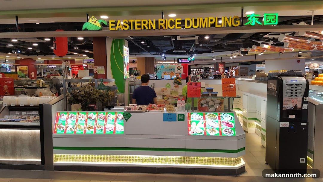 A food stall with a sign that reads "EASTERN RICE DUMPLING" and Chinese characters. The stall displays various food items in refrigerated cases, including dumplings and other dishes. There are also drinks and a coffee machine.