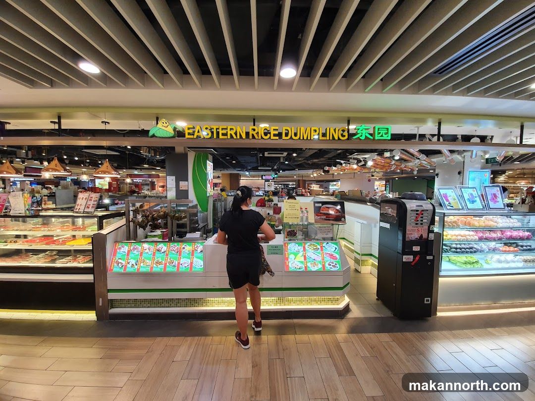 A woman stands in front of a food stall with a sign that reads "EASTERN RICE DUMPLING" and Chinese characters. The stall displays various food items in refrigerated cases and on illuminated menu boards. To the right, a coffee machine is visible. The flooring is made of wood-like tiles.