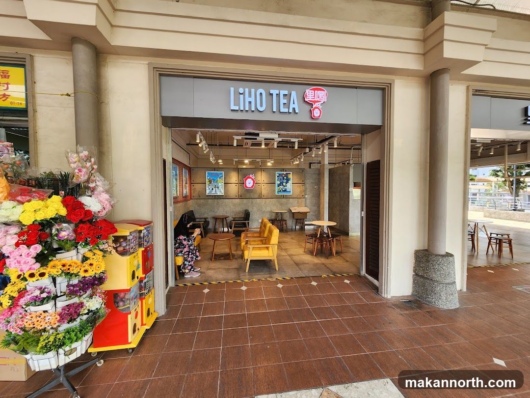 The exterior of a Liho Tea shop. The shop has a modern design with a gray facade and a prominent "LIHO TEA" sign above the entrance. Inside, there are tables and chairs for customers, and some artwork on the walls. To the left of the shop entrance, there is a display of artificial flowers and a gumball machine.