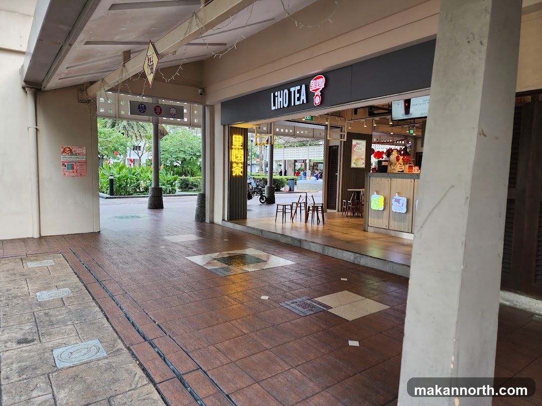 The exterior of a Liho Tea shop. The shop has a prominent sign with the brand name and logo. Inside, tables and chairs are visible, along with some decorative items. The entrance is open, revealing the interior of the shop.