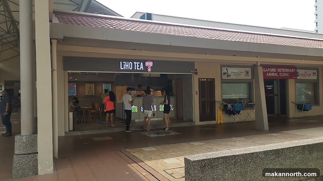 A row of shops with a prominent "LI HO TEA" sign above one entrance. People are seen inside and queuing outside the tea shop. To the right, a sign reads "SINGAPORE VETERINARY ANIMAL CLINIC" with logos for "Royal Canin" and another veterinary service. Clothes racks with laundry are placed outside the clinic.