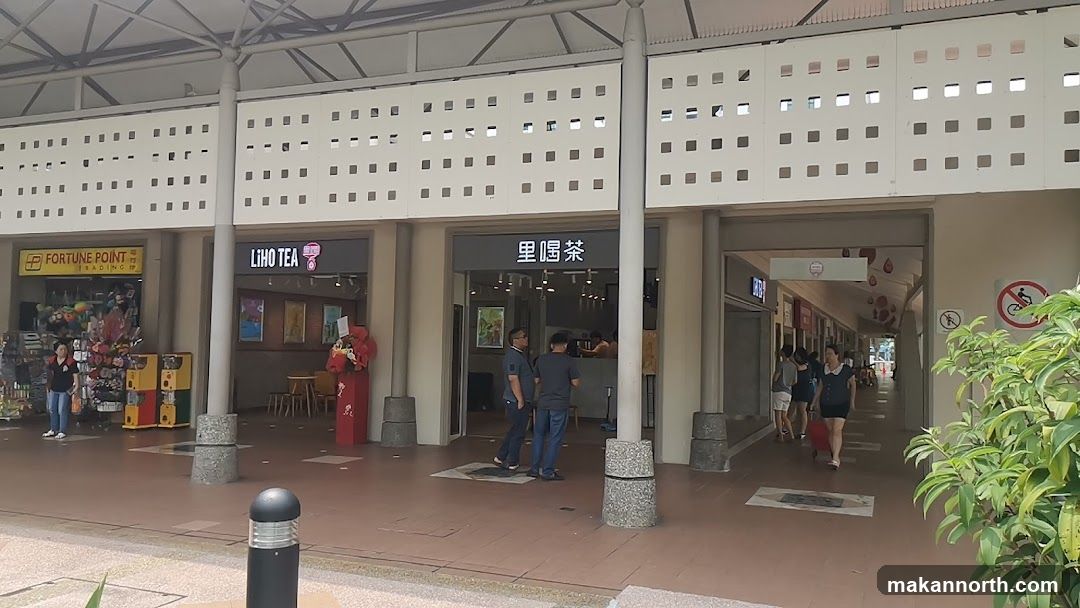 A row of storefronts under a covered walkway. On the left is a store with a yellow sign that says "FORTUNE POINT TRADING". In the center is a store with a dark sign that says "LIHO TEA". To the right of Liho Tea is another store with Chinese characters above the entrance. People are seen walking in the walkway and entering the stores.
