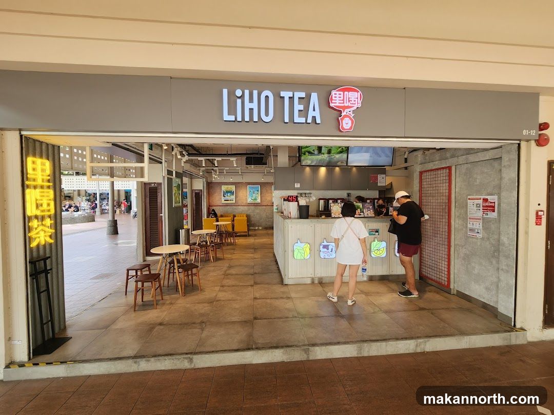 The exterior of a LiHO TEA shop. The shop has a large sign with the name "LIHO TEA" and a logo. There are tables and chairs outside the shop. Inside, there is a counter with staff and customers.