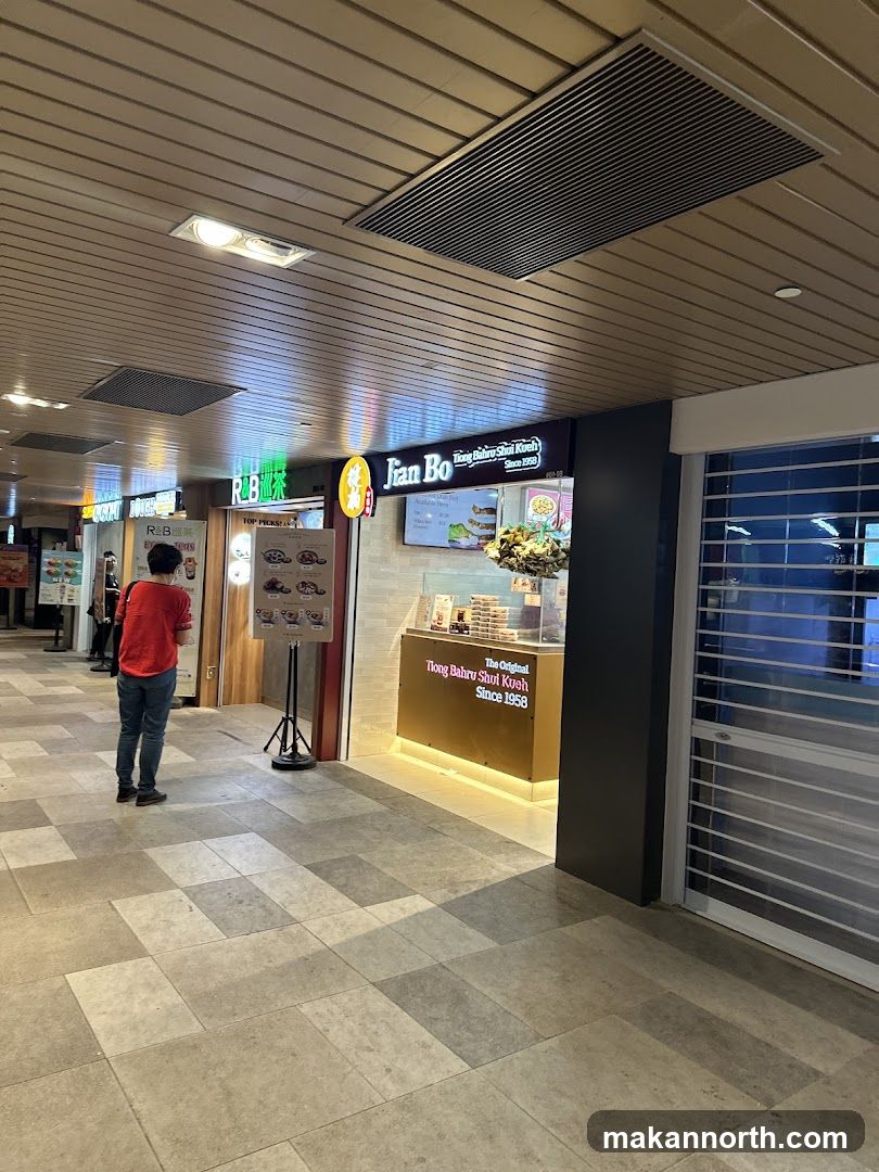 A shop front with a sign that reads "Jian Bo The Original Tiong Bahru Shui Kueh Since 1958". There is a display case with food items and a person standing in front of the shop.