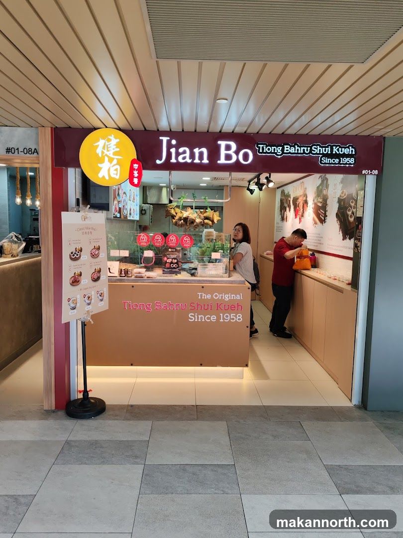 A storefront for Jian Bo Tiong Bahru Shui Kueh, a business established in 1958. The shop features a prominent sign with the business name and logo. Inside, there are displays of food items and customers interacting with staff. A menu board is visible to the left of the entrance.