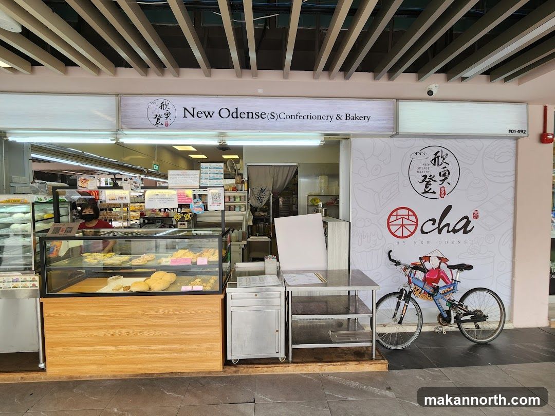 The storefront of a bakery and confectionery shop. A display case in front shows various baked goods. A sign above the entrance reads "New Odense (S) Confectionery & Bakery". A large graphic on the right side of the storefront features the text "cha BY NEW ODENSE" and an illustration of a bicycle with a figure.
