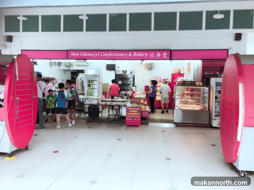 A wide shot of the exterior of a confectionery and bakery. The shop's name is displayed prominently on a pink banner above the entrance. Inside, various baked goods and pastries are displayed on tables and in glass cases. Several customers and staff members are visible.