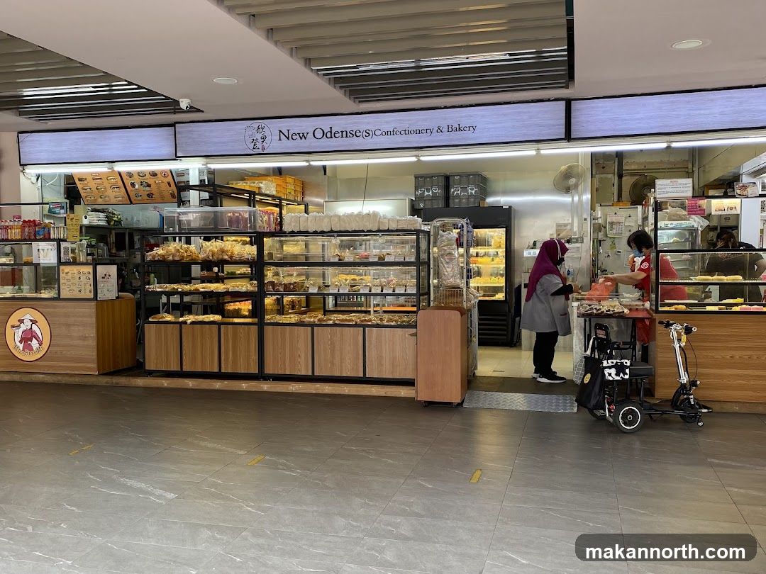 The exterior of a confectionery and bakery shop. Display cases filled with baked goods are visible, along with a counter where two staff members are serving customers. A sign above the entrance reads "New Odense(S) Confectionery & Bakery".