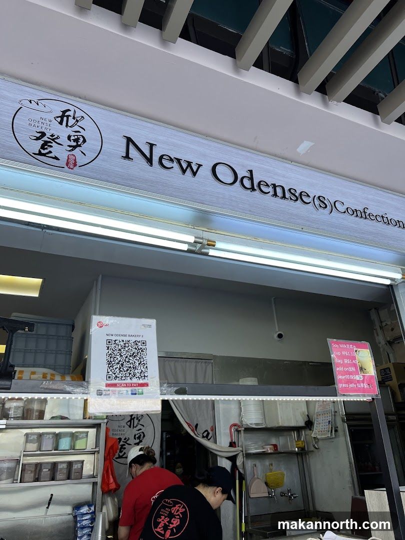The exterior of a bakery with a prominent sign displaying "New Odense (S) Confectionery" and a logo. Below the sign, a QR code for payment is visible, along with a small menu board showing drink options and prices. The interior of the shop, including staff and display shelves, is partially visible.