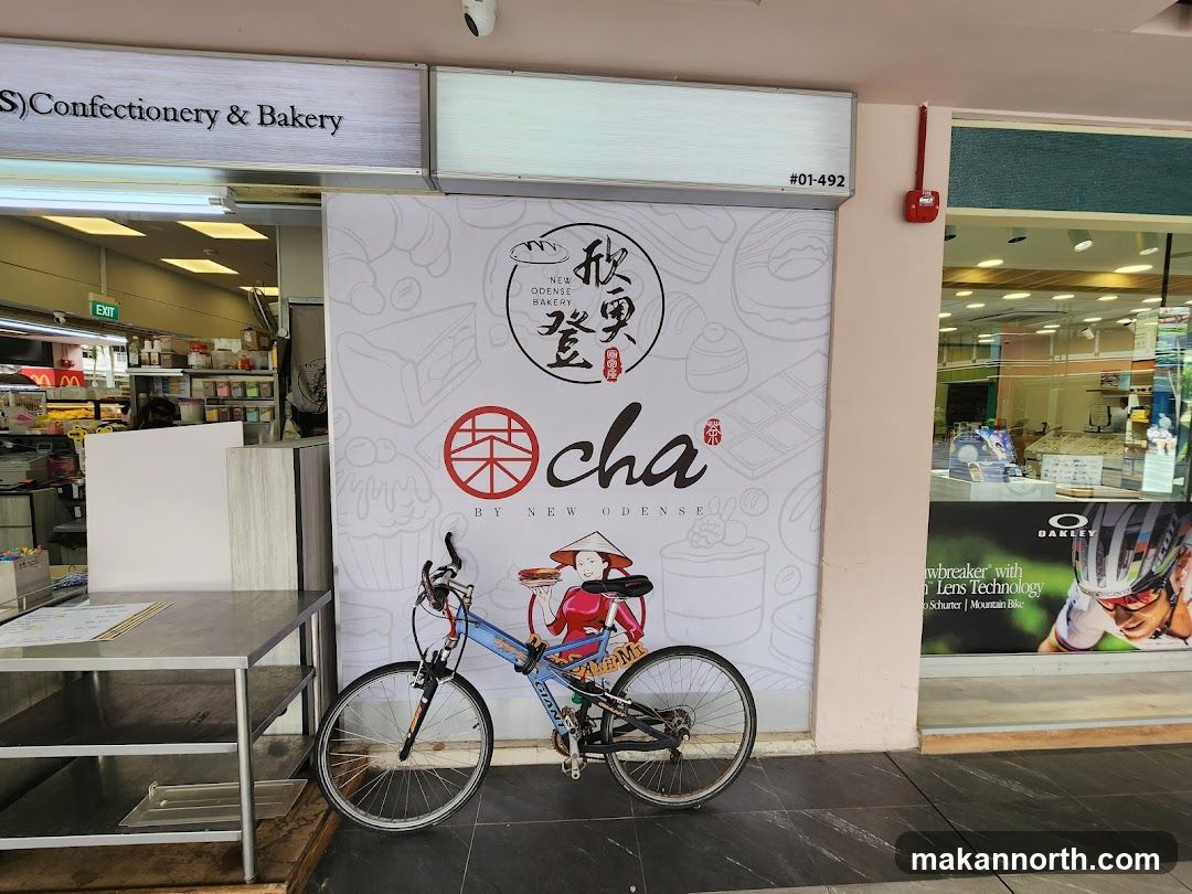 The exterior of a confectionery and bakery shop. A large graphic on the wall features the text "NEW ODENSE BAKERY" and "cha BY NEW ODENSE". A bicycle is parked in front of the graphic. To the left, an entrance with an "EXIT" sign is visible, showing shelves stocked with items inside. To the right, a shop window displays promotional material for Oakley eyewear and mountain biking.