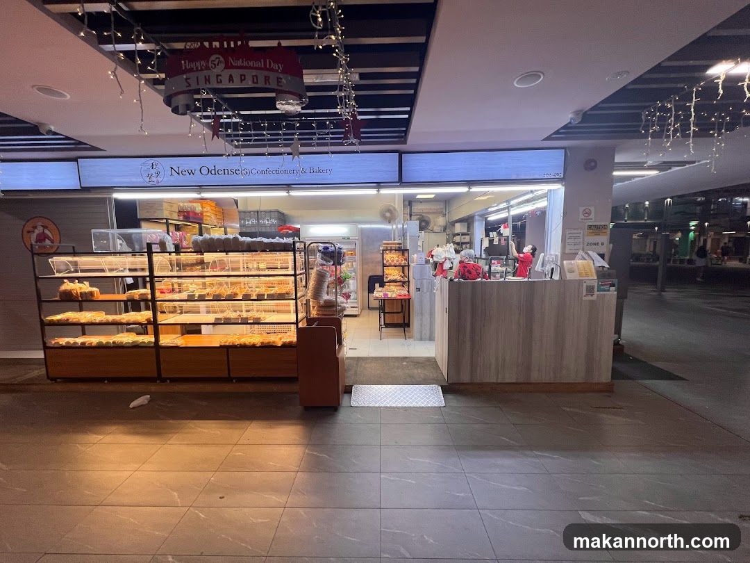 The exterior of a confectionery and bakery shop. Display shelves filled with various baked goods are visible in front of the store. The shop's name, "New Odense Confectionery & Bakery," is displayed prominently on a sign above the entrance. Decorations for National Day are also present.