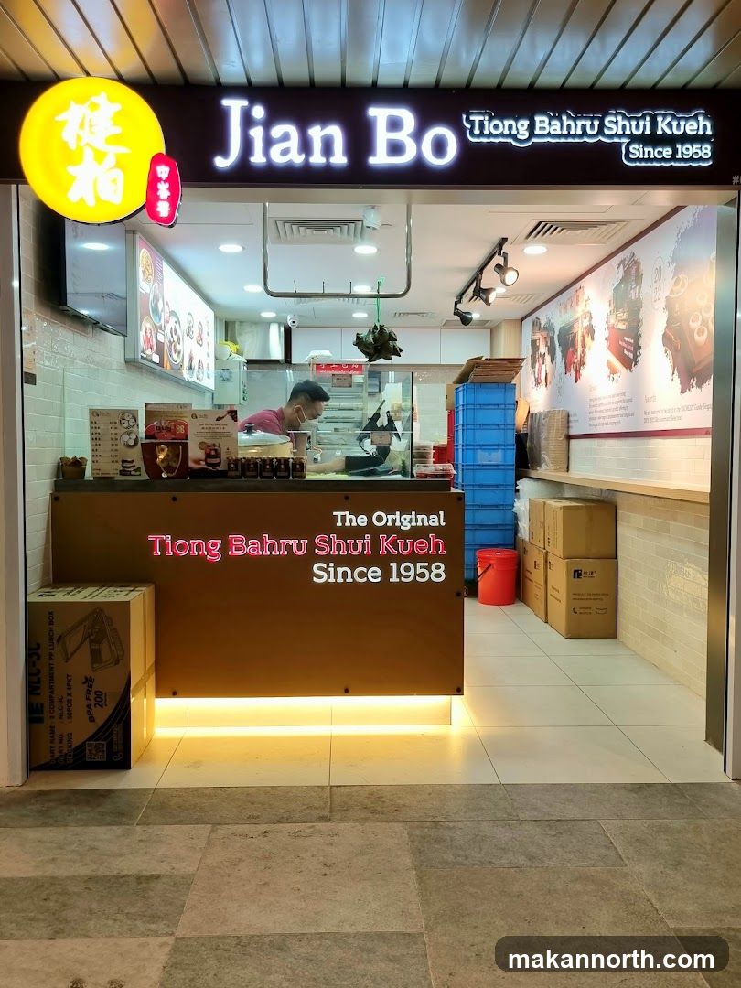 The exterior of a food stall with a prominent sign reading "Jian Bo Tiong Bahru Shui Kueh Since 1958". A person is visible behind the counter preparing food. There are boxes and other items stacked around the stall.