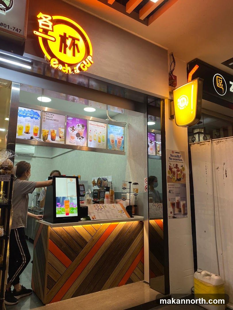 The exterior of a bubble tea shop called Each a Cup. A sign with the shop's logo is visible above the entrance. Inside, staff are preparing drinks behind a counter. A menu board displays various drink options.