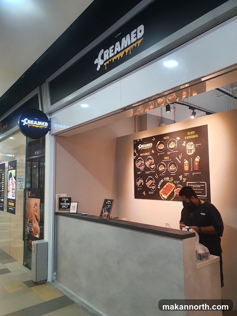 A view of the exterior of a shop called Creamed. The shop has a prominent sign above the entrance and a circular illuminated sign on the left. Inside, a menu board displays food items. A staff member is visible behind the counter.