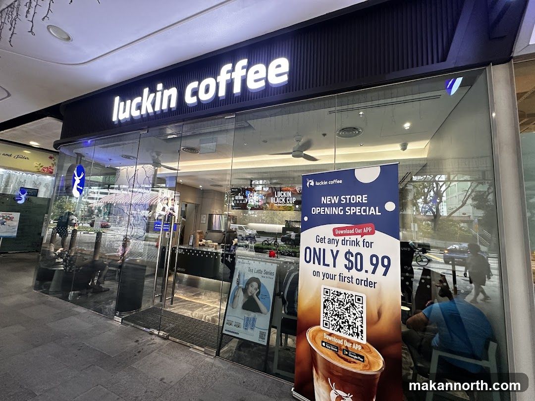 The exterior of a Luckin Coffee shop with large glass windows and doors. The prominent "luckin coffee" sign is illuminated above the entrance. Inside, a counter and seating area are visible. A large banner advertises a "New Store Opening Special" offering any drink for $0.99 on the first order with a QR code for app download. Another poster displays "Tea Latte Series".