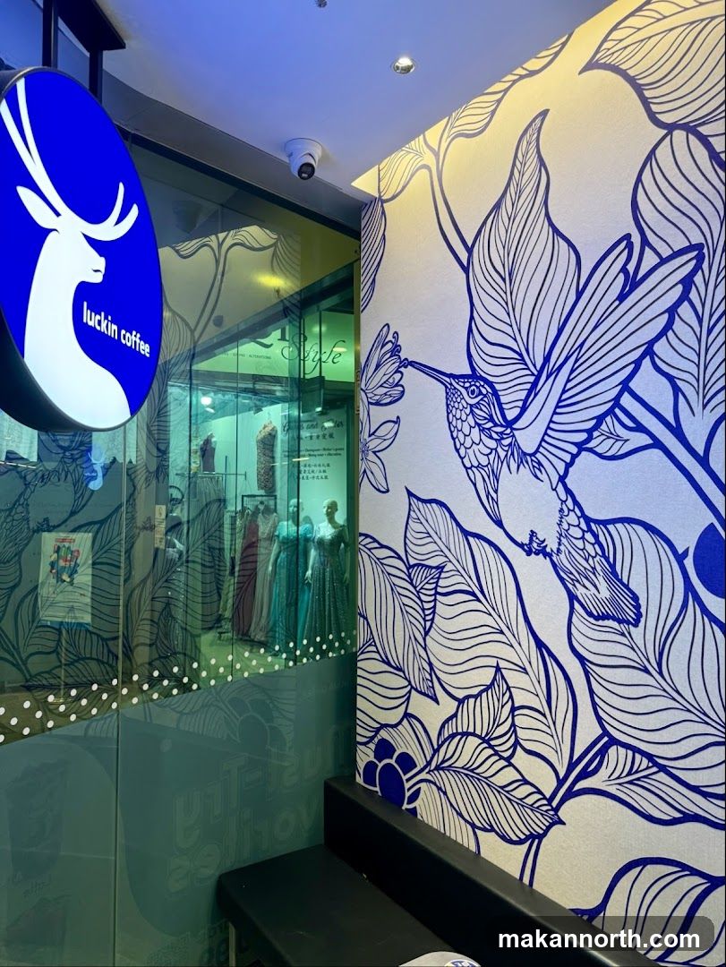 A blue and white sign with a deer logo and the text "luckin coffee" is visible on the left side of the image. The sign is illuminated and oval-shaped. The storefront features large glass windows, through which mannequins dressed in formal wear can be seen. The interior of the shop is dimly lit. A wall inside the shop is decorated with a large blue line drawing of a hummingbird and foliage. A black bench is positioned in front of the decorated wall.
