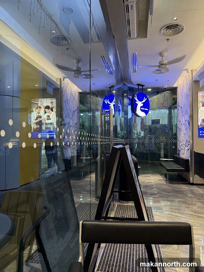 A view through the glass doors of a Luckin Coffee shop. The interior features blue and white decor with a deer logo. An A-frame sign is placed outside the entrance.