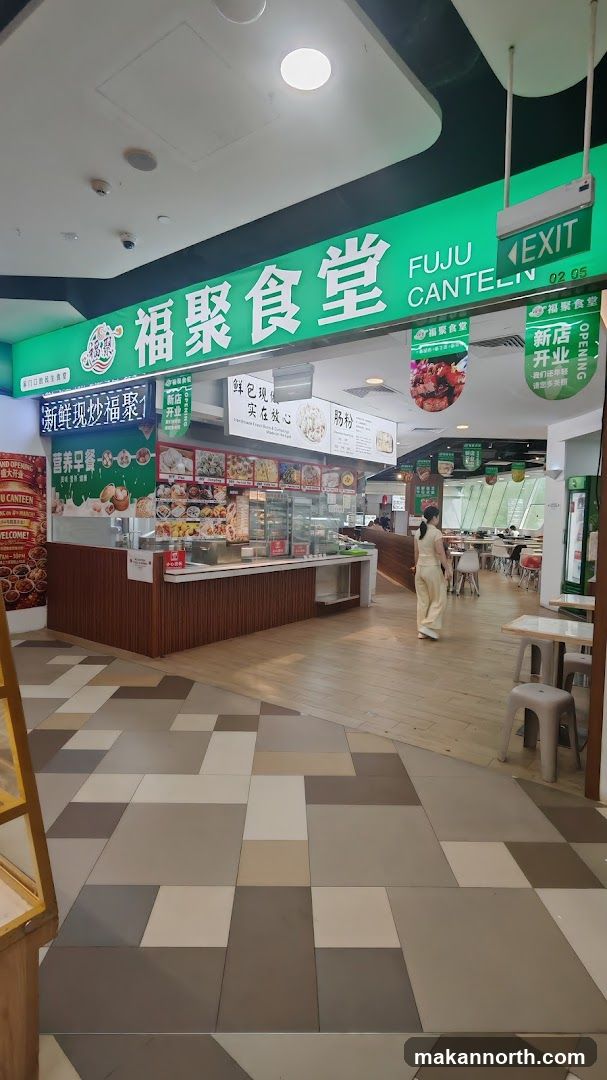 The exterior of a food stall with a large green sign that reads "FUJU CANTEEN" in English and Chinese characters. There are various food displays and menus visible.