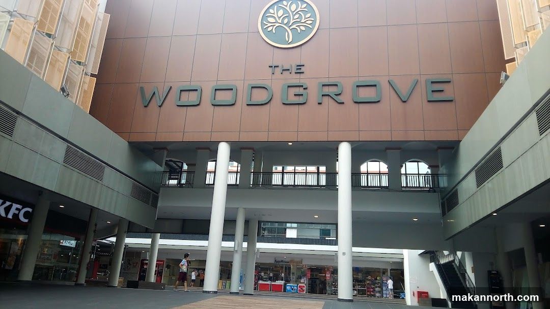 A wide shot of the exterior of The Woodgrove shopping mall. The mall's name is prominently displayed in large, dark green letters on a brown facade, with a circular logo above it featuring a stylized tree. Below the main facade, there are multiple levels with shops visible, including a KFC on the left. White pillars support the upper levels, and a walkway with railings is visible on the second floor. People are seen walking in the open area in front of the shops.