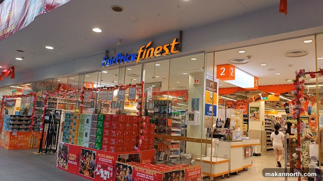 The exterior of a FairPrice Finest supermarket. The store has large glass windows and doors, with the store's name prominently displayed above the entrance. Inside, shelves are stocked with various products, and there are displays of beverages and other items. Decorations for Chinese New Year are visible, including red and pink floral arrangements and banners.