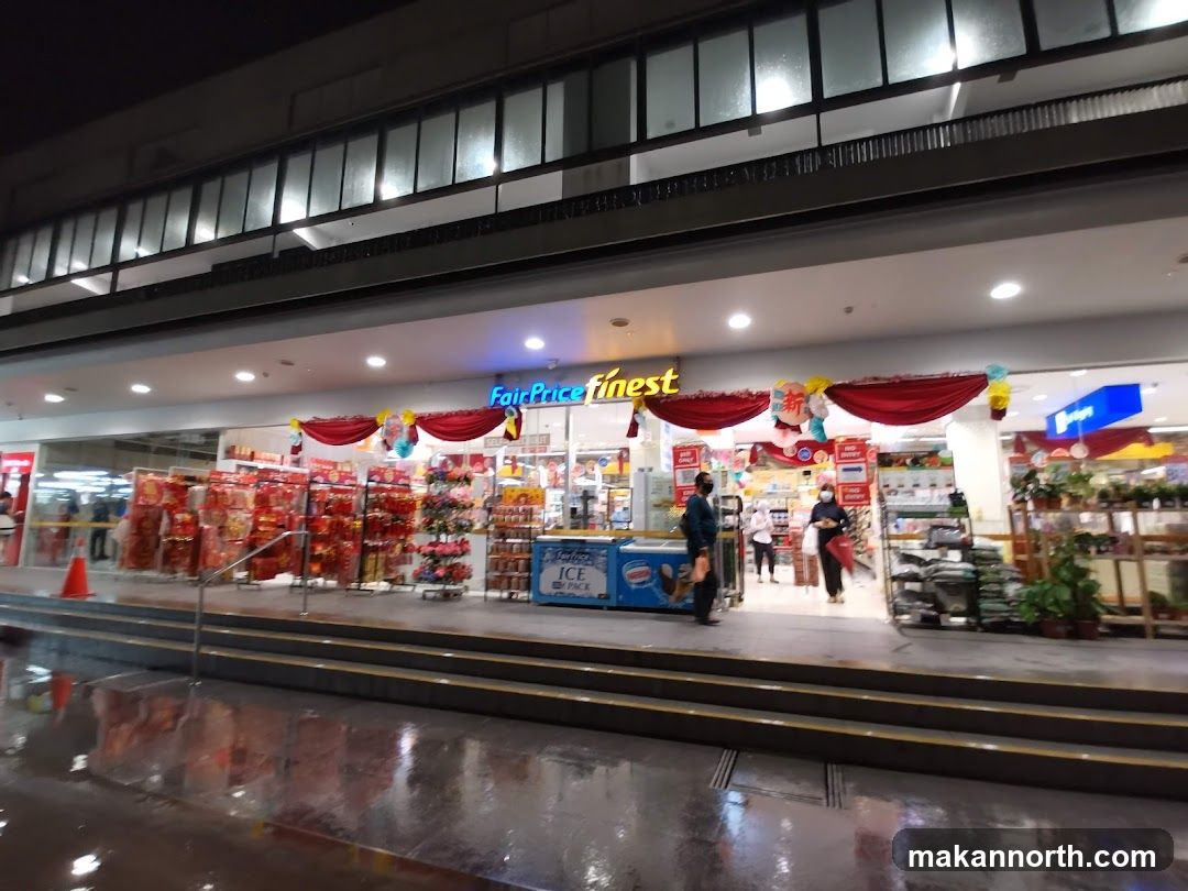 The exterior of a FairPrice Finest supermarket at night. The entrance is wide and well-lit, with displays of red festive items and flowers visible inside. Red drapes and balloons decorate the entrance. Several people are seen entering and exiting the store. The store's signage "FairPrice Finest" is prominently displayed above the entrance.