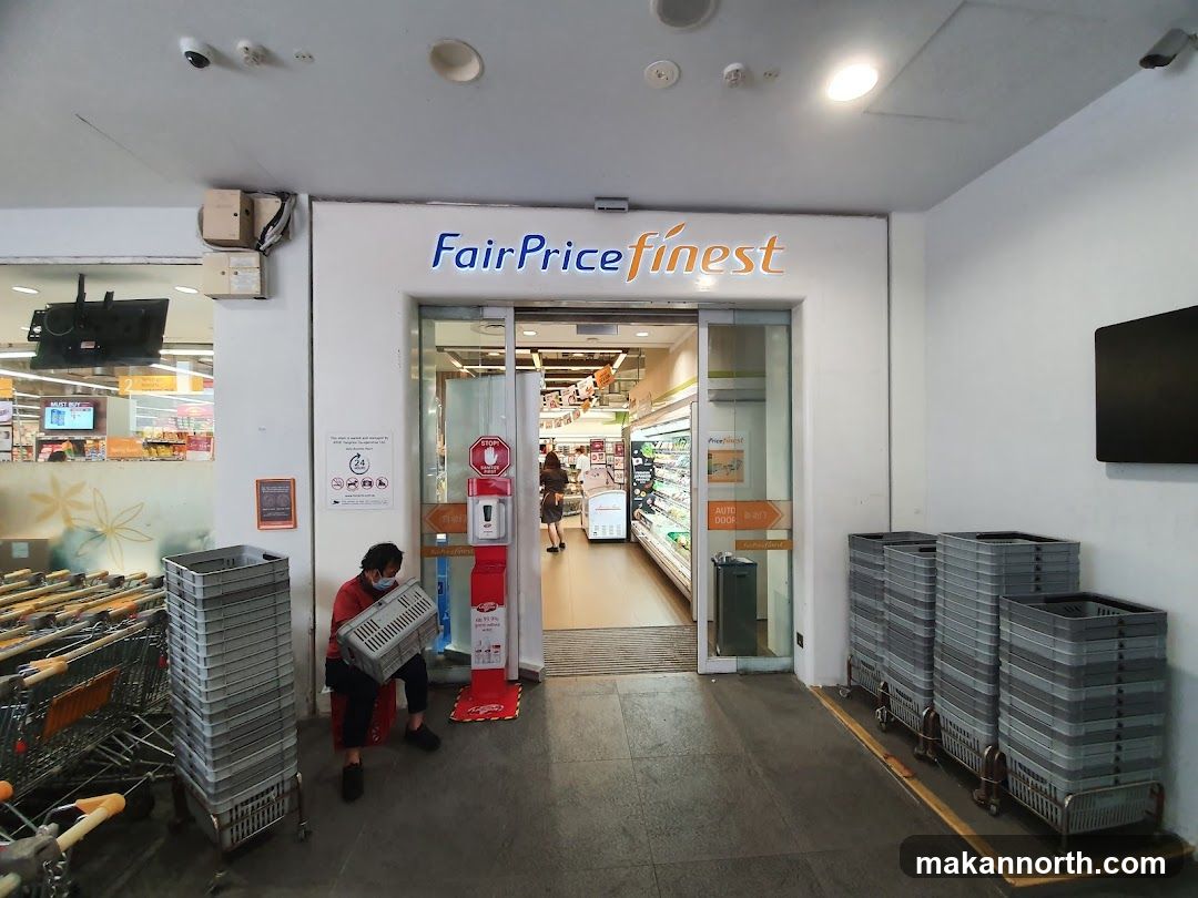 The entrance to a FairPrice Finest supermarket. The store's name is displayed in blue and orange lettering above the automatic glass doors. To the left of the entrance, shopping carts are stacked, and a person is sitting with a basket. To the right, more shopping baskets are stacked. Inside the store, aisles of products are visible.