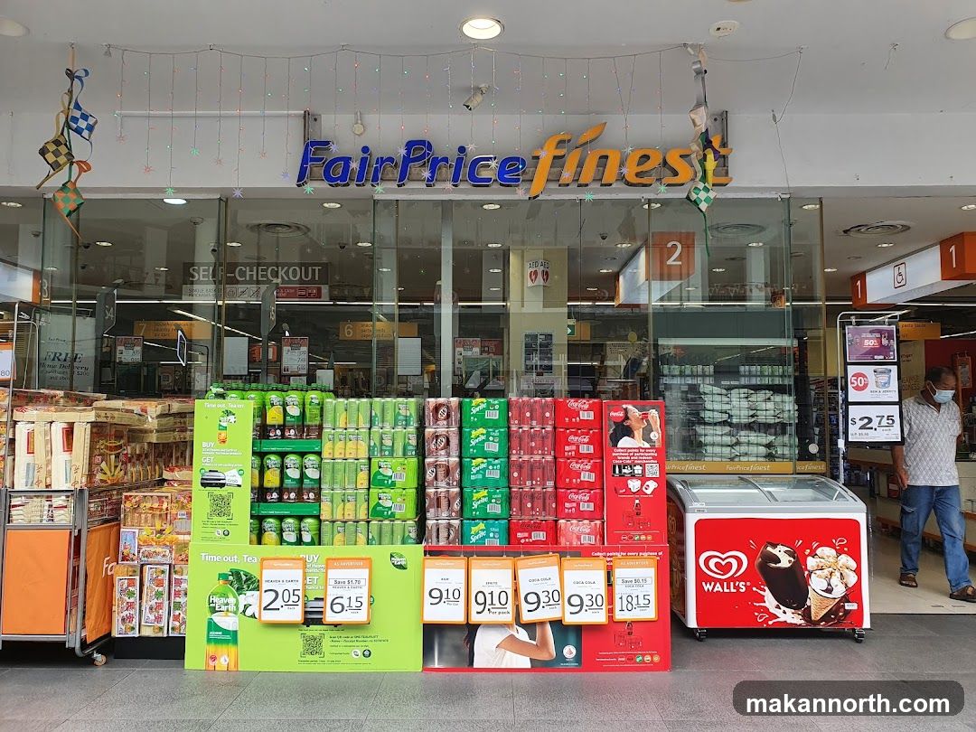The exterior of a FairPrice Finest supermarket with a prominent sign above the entrance. Displays of beverages and ice cream are set up outside the store. Decorations, possibly for a holiday, hang above the entrance. A person is visible walking near the store.