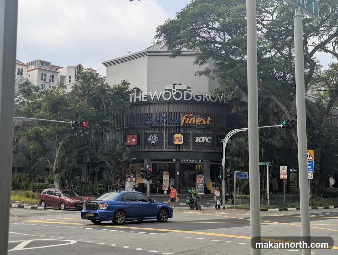 The exterior of The Wood Grove building, a commercial establishment with multiple storefronts. Prominent signage includes "THE WOODGROVE" and "FairPrice finest". Several fast-food chains like McDonald's, Burger King, and KFC are visible, along with other businesses indicated by their logos. The building has a modern architectural style with dark paneling and large windows. Trees surround the structure, and a street with traffic lights and cars is in the foreground.