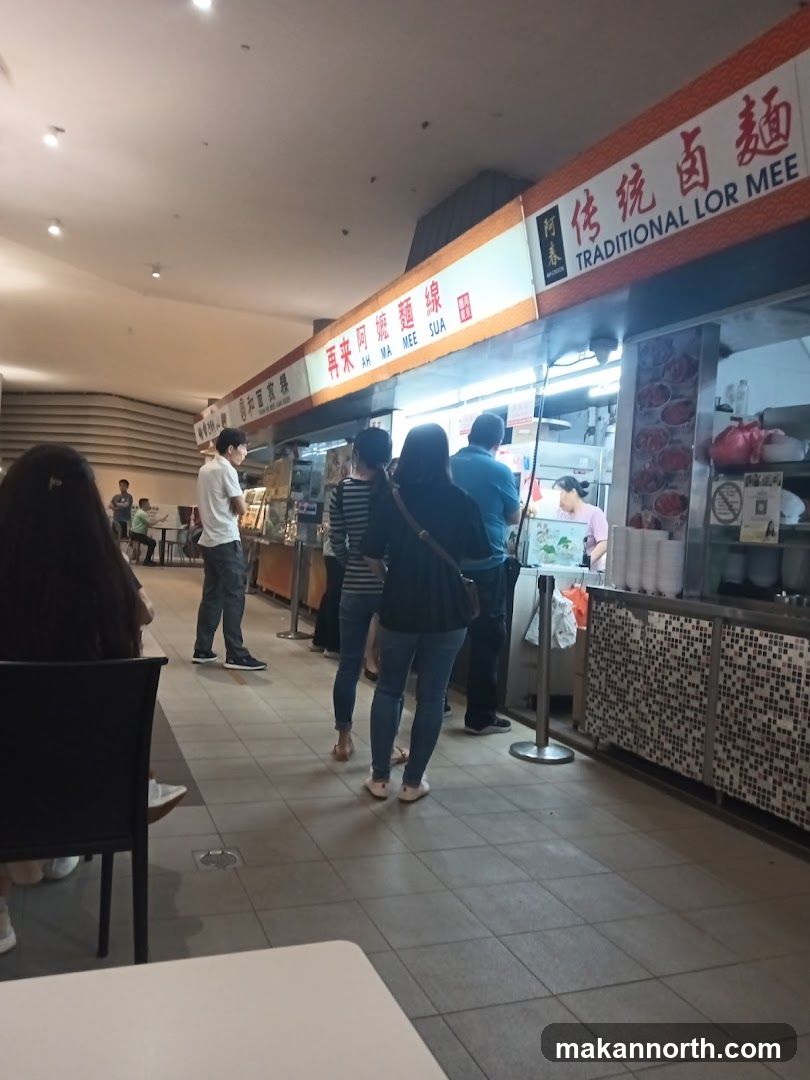 A food stall with a sign that reads "Traditional Lor Mee" in English and Chinese characters. Several people are standing in line to order food. The stall appears to be in a food court or hawker center.