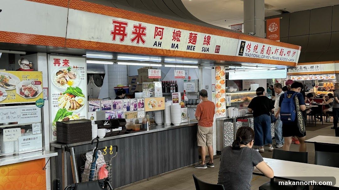 A food stall with a prominent sign displaying "AH MA MEE SUA" in English and Chinese characters. The stall offers traditional Lor Mee, Prawn Noodle, and Laksa. There are menu boards with pictures of dishes and prices. Customers are seen ordering and waiting for their food.