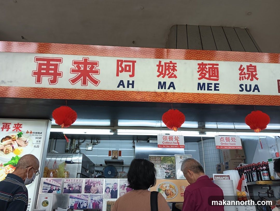 A food stall with a large sign displaying the name "AH MA MEE SUA" in both Chinese and English. Red lanterns hang above the counter. There are menus and pictures of food displayed. People are seen queuing and ordering.