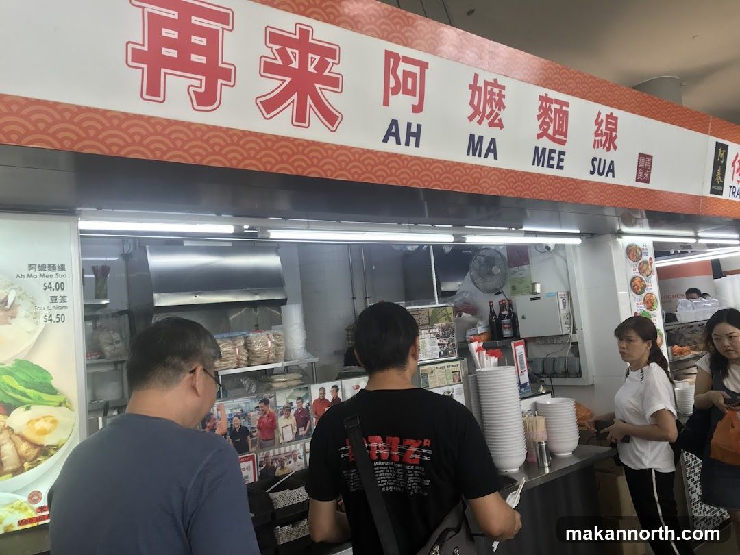 A food stall with a prominent sign displaying "AH MA MEE SUA" in both Chinese and English. The stall appears to be busy with customers. A menu board is visible on the left, listing "Ah Ma Mee Sua" for $4.00 and "Tau Chiam" for $4.50.