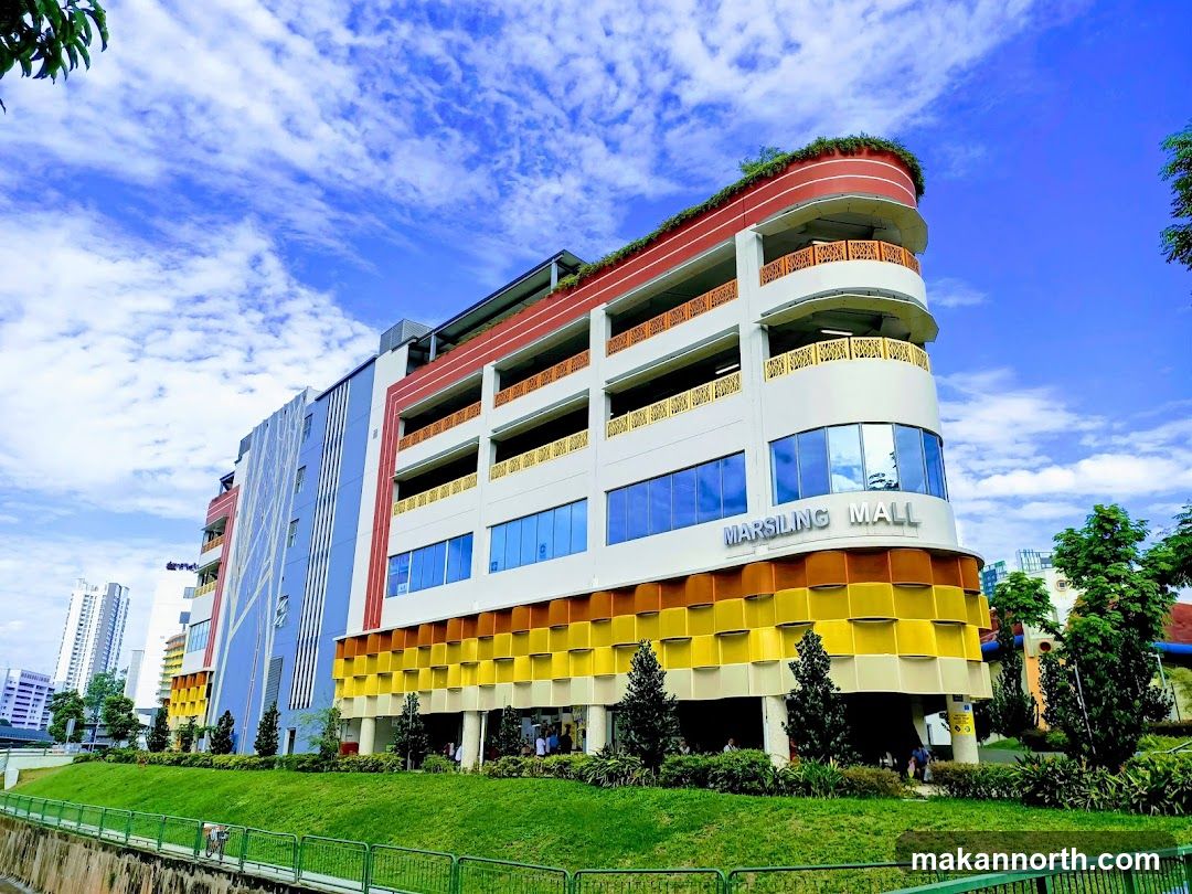 A multi-story building with a colorful facade, featuring a prominent sign that reads "MARSILING MALL". The building has a modern architectural style with balconies, large windows, and a green roof. It is surrounded by lush greenery and a bright blue sky with scattered clouds.