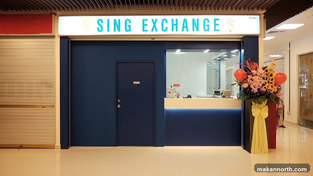 The exterior of a Sing Exchange shop. The shop has a blue facade with a large illuminated sign above that reads "SING EXCHANGE" with dollar signs on either side. To the left of the entrance is a closed metal shutter. To the right of the entrance is a counter with a window, and a large floral arrangement with balloons. A sign on the door reads "Staff Only". A "Closed" sign is visible on the counter window.