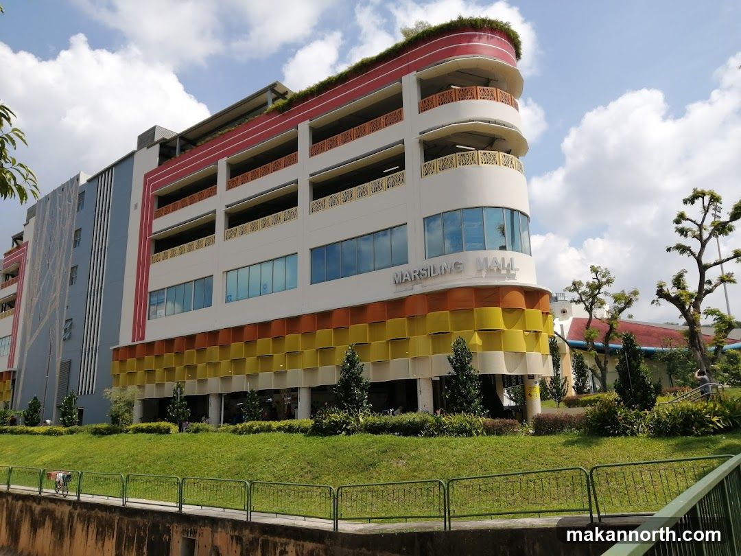 A multi-story building with a curved facade, featuring a colorful design with bands of orange, yellow, and white. The words "MARSILING MALL" are visible on the building. The structure has multiple levels with balconies and windows, and is surrounded by greenery and trees. The sky above is partly cloudy.