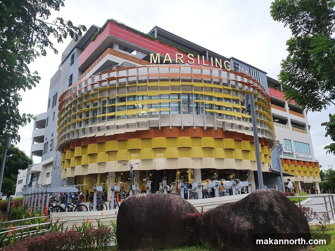 A multi-story building with a distinctive curved facade featuring yellow, orange, and white panels. The name "MARSILING MALL" is visible in large letters at the top of the building. A Burger King sign is also visible on the right side of the building. In front of the building, there is a bicycle parking area with several bicycles. Large rocks and landscaping are in the foreground.