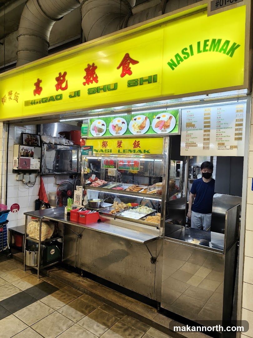 A food stall with a bright yellow sign displaying Chinese characters and "GAO JI SHU SHI" in English, along with "NASI LEMAK" in green. Below the main sign, there is another sign with images of nasi lemak dishes and the text "NASI LEMAK" in both Chinese and English. The stall features a display counter with various food items and a person wearing a mask standing behind it. A menu is visible on the right side of the stall.