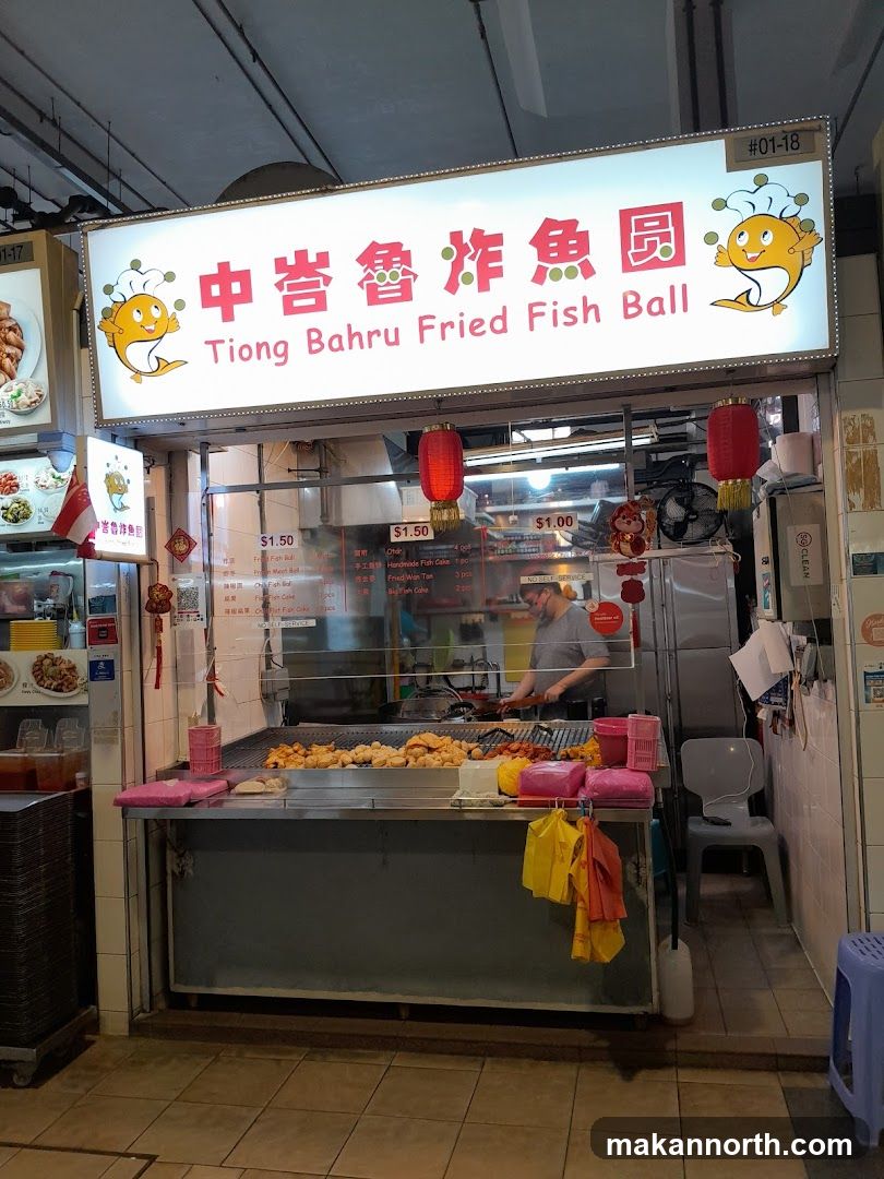 A food stall with a large illuminated sign displaying the name "Tiong Bahru Fried Fish Ball" in English and Chinese. The stall features a display of fried food items and a person cooking behind the counter. Red lanterns hang above the counter.