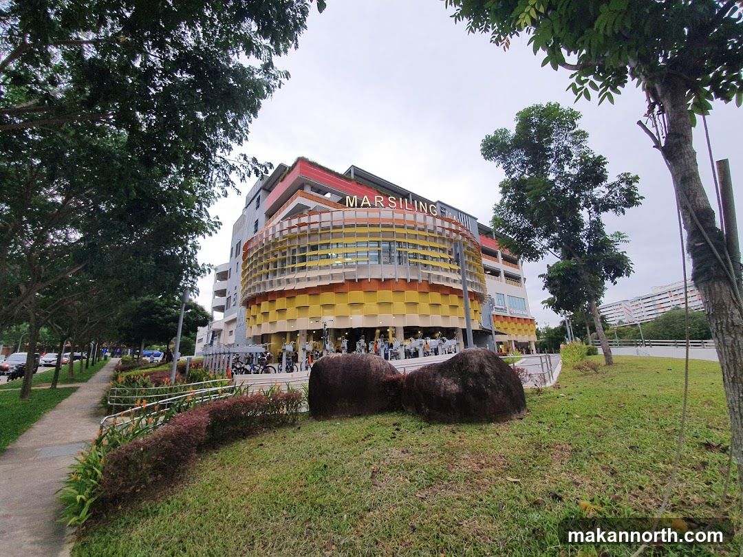 A wide shot of Marsiling Mall, a multi-story building with a distinctive curved facade featuring yellow, orange, and white panels. The name "MARSILING" is visible in red lettering at the top of the building. The mall is surrounded by trees and greenery, with a sidewalk and road in the foreground.